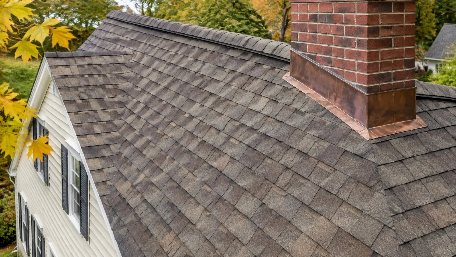 Close view of new brown dimensional shingles meeting a brick chimney with copper flashing, a yellow maple leaf at the edge