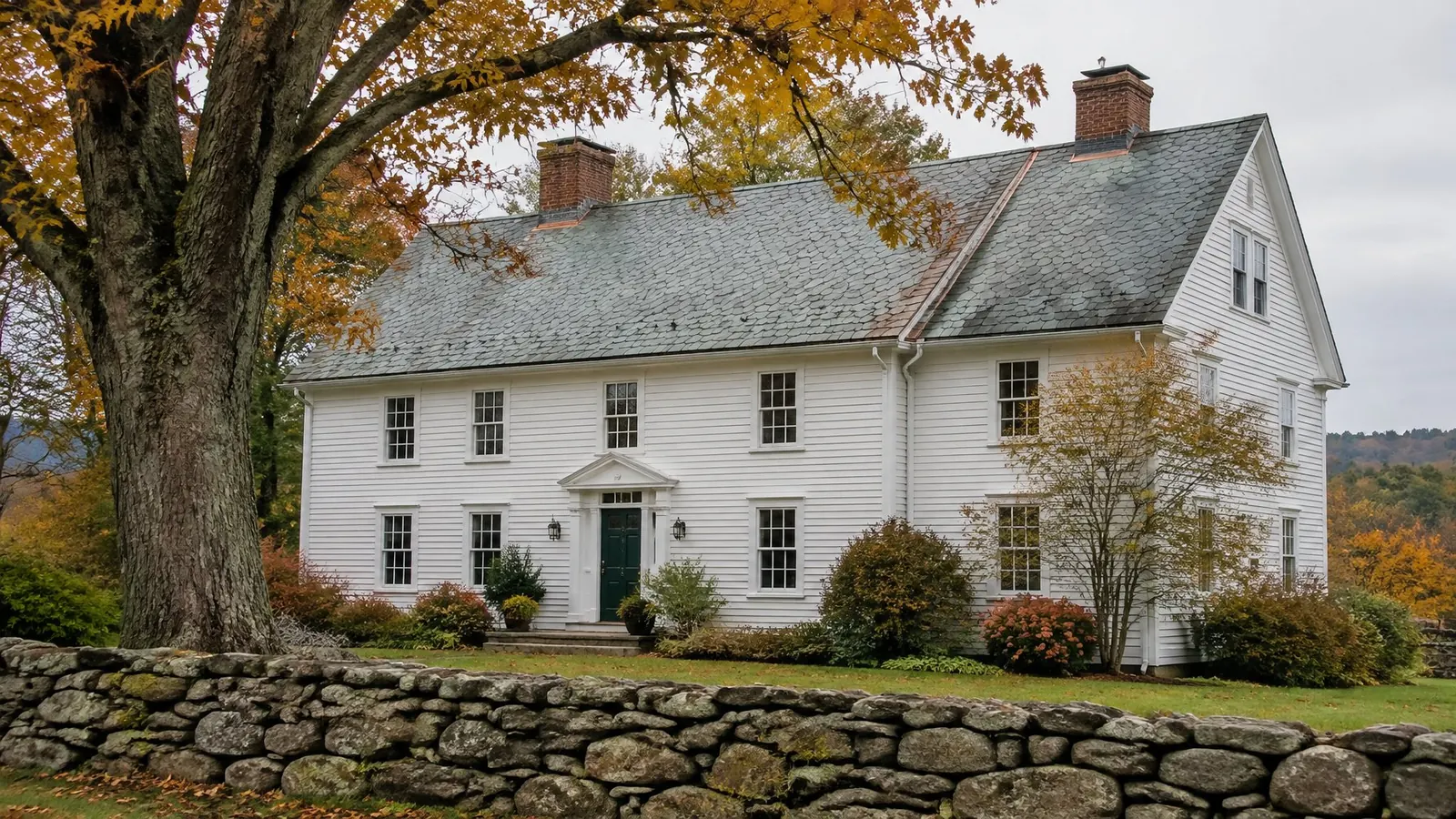 White clapboard colonial with a gray-green slate roof, copper ridge and valley flashing, a large maple and a fieldstone wall